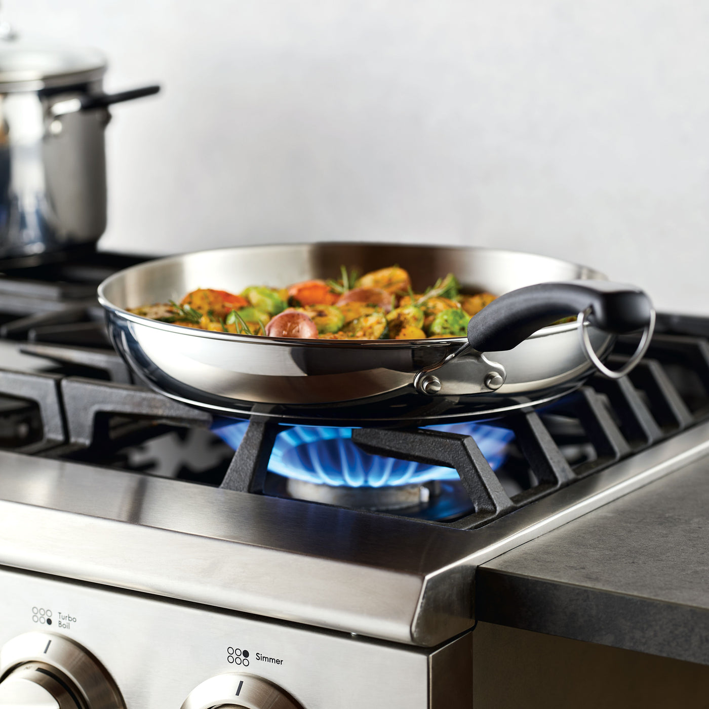 A 12.25-Inch Stainless Steel Frying Pan rests on a gas stove, cooking food above a vibrant blue flame.