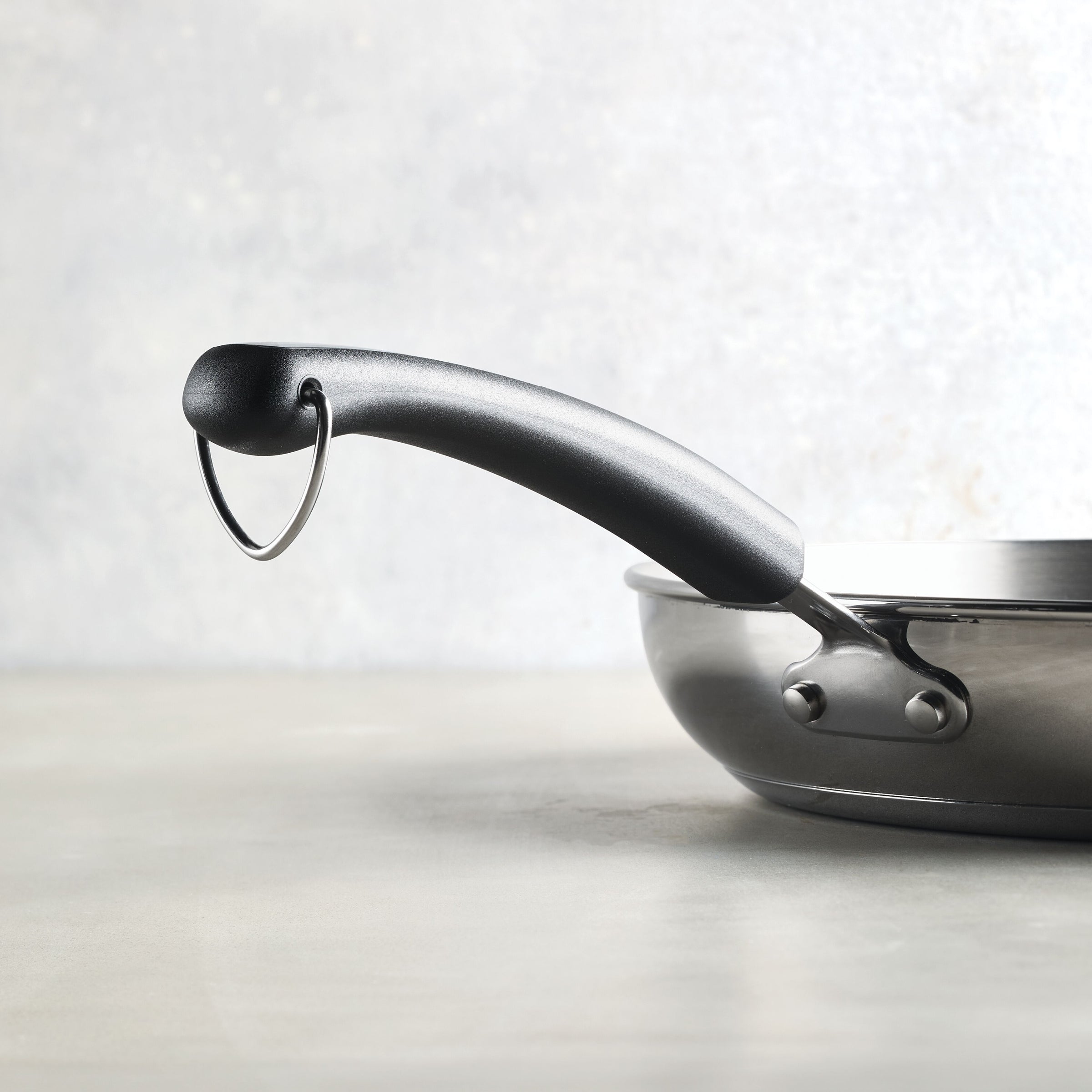 Close-up of a black pan handle on a gray background, highlighting the sleek design of the 12.25-Inch Stainless Steel Frying Pan from Farberware Brilliance cookware.