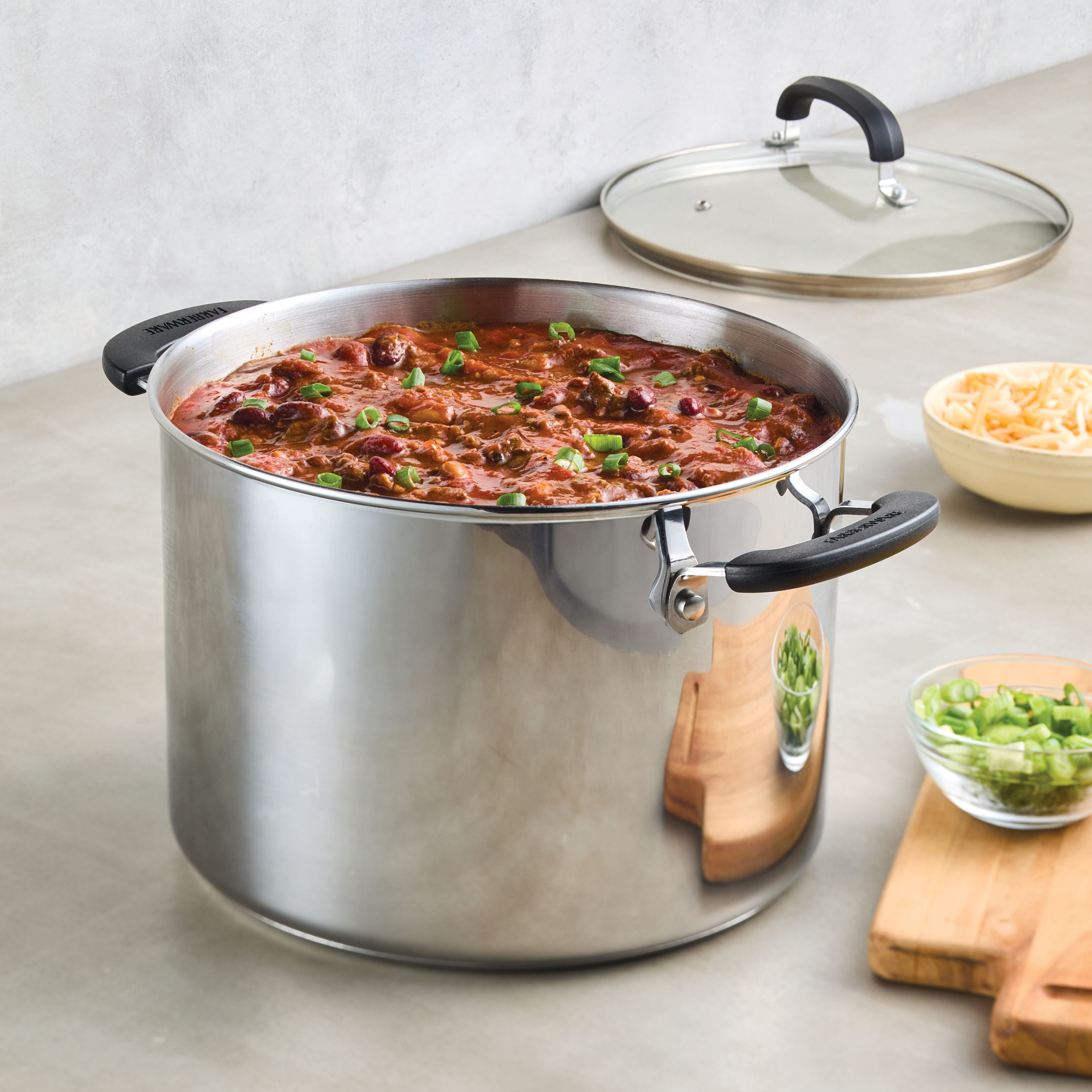 An inviting pot of chili in an 8-Quart Stainless Steel Stockpot from Farberware Brilliance, adorned with beans and garnish, sits enticingly beside shredded cheese and fresh green onions.