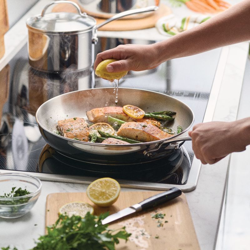 A hand squeezes lemon over salmon in a Stainless Steel 12.25-Inch Frying Pan.