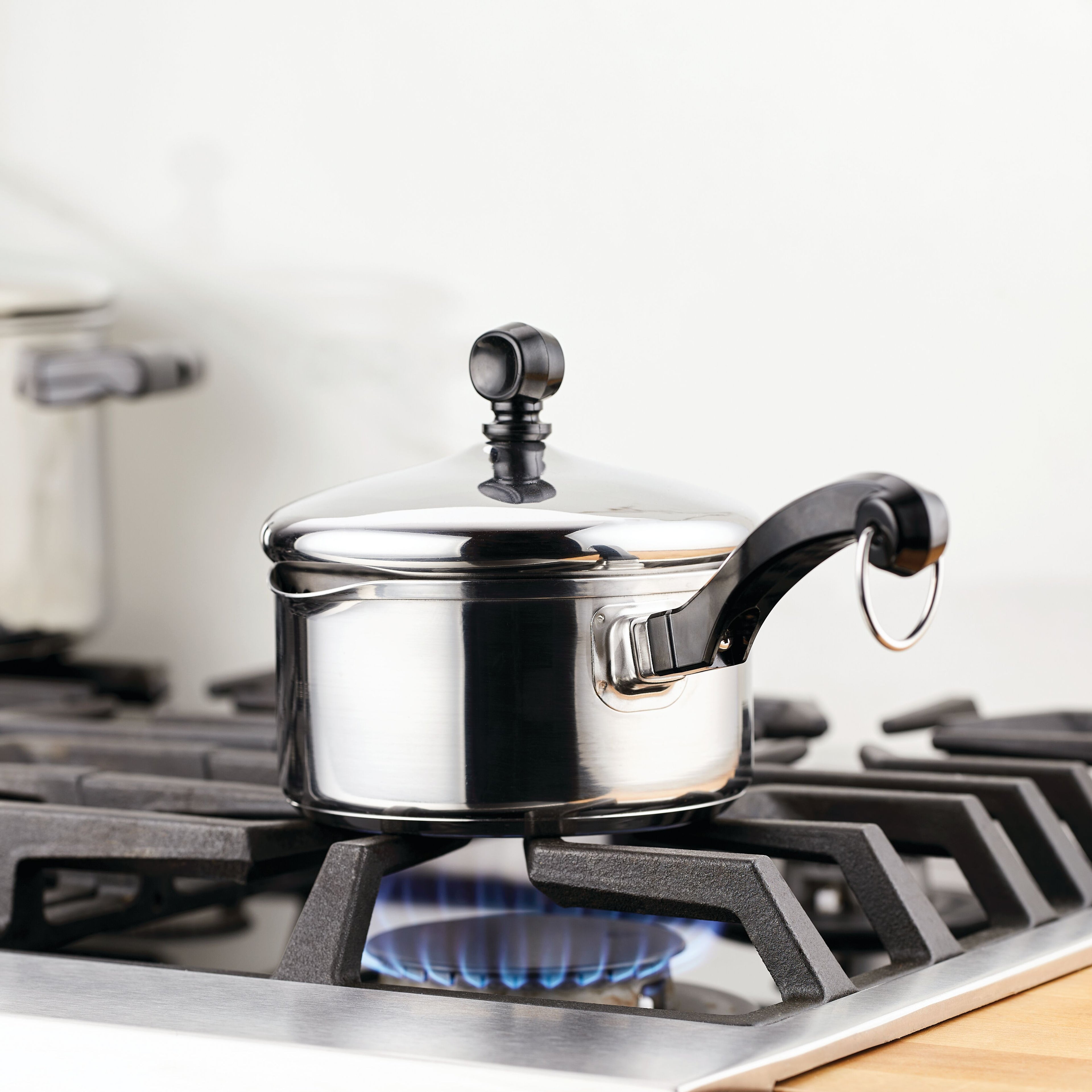 The Classic Series Stainless Steel Saucepan With Straining Lid sits on a lit gas stove burner, making it perfect for preparing steamed veggies in your kitchen.