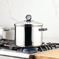 An 8-quart Classic Series Stainless Steel Stockpot with a straining lid sits on a gas stove burner, ideal for large-scale cooking in the kitchen.