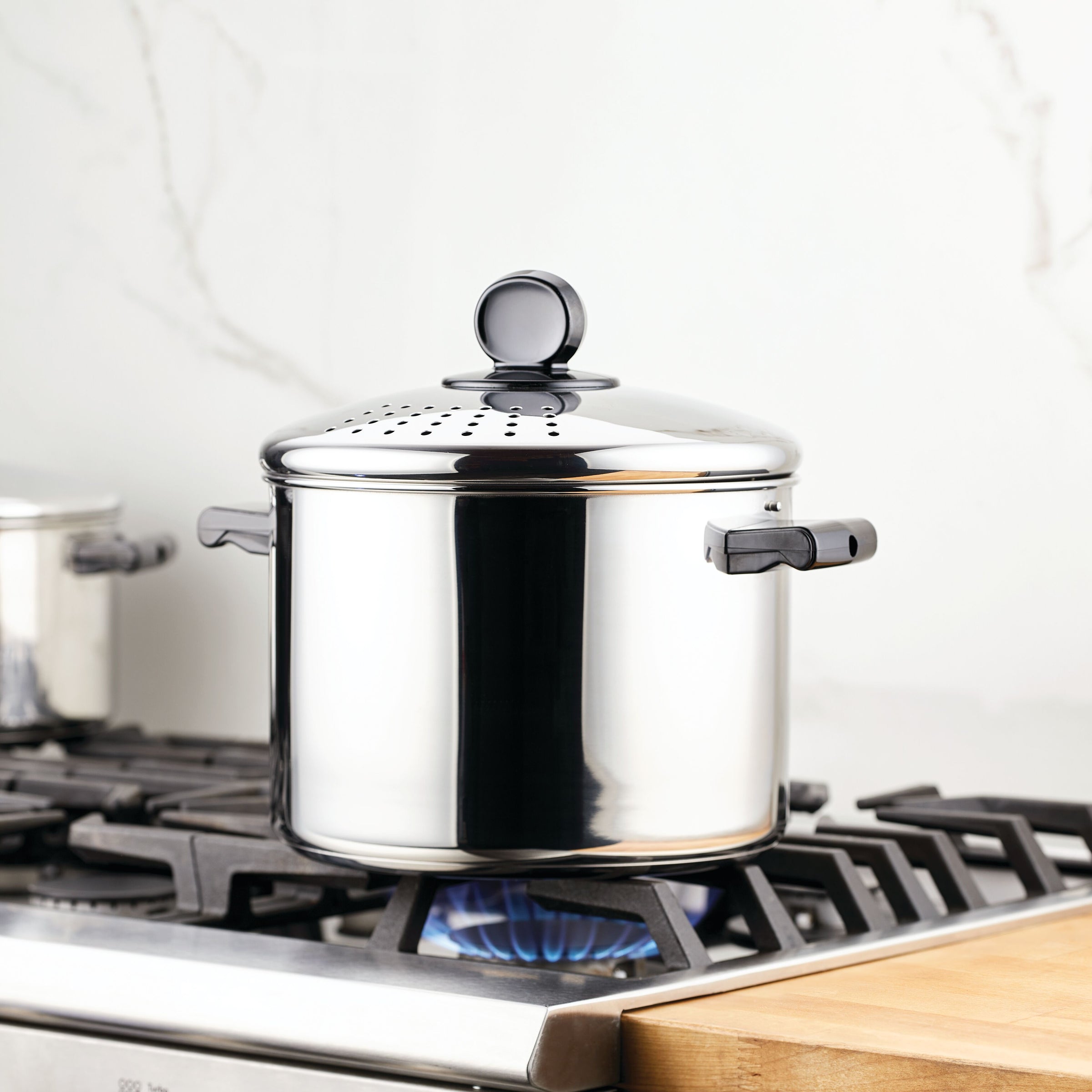 An 8-quart Classic Series Stainless Steel Stockpot with a straining lid sits on a gas stove burner, ideal for large-scale cooking in the kitchen.