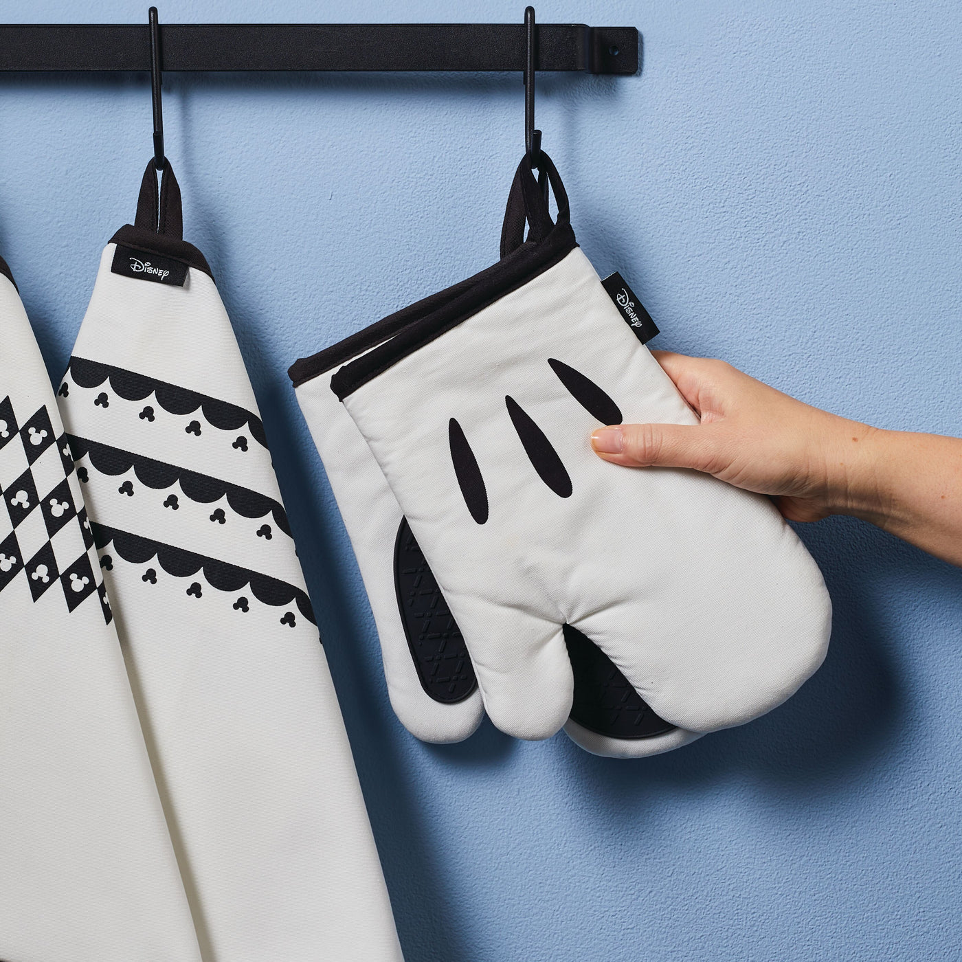 A hand holds an oven mitt from the Monochrome 5-Piece Set against a blue wall, adding a touch of Disney Home charm to your kitchen set.
