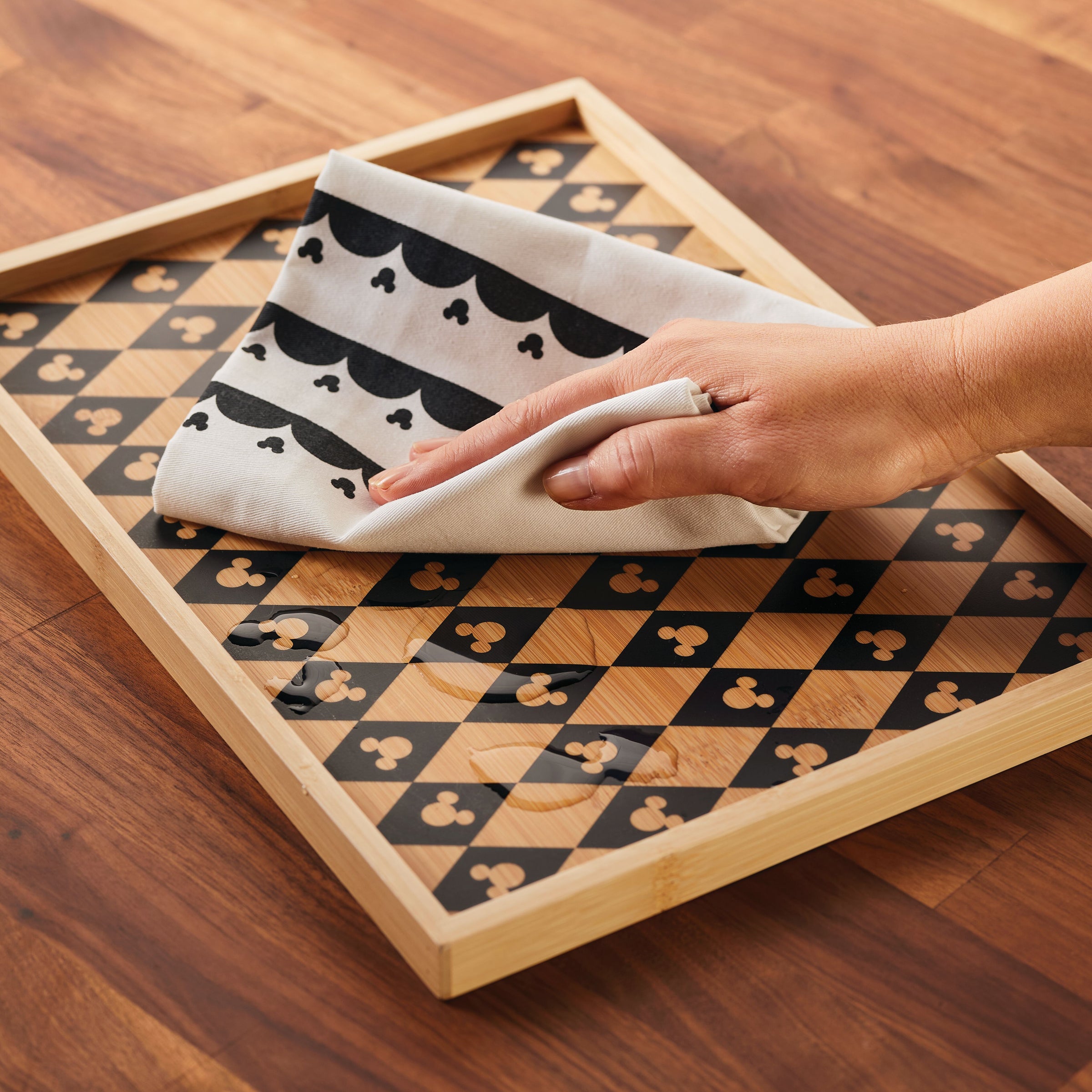 A hand wipes spills on a wooden tray using kitchen towels from Disney Home Monochrome, featuring subtle Mickey Mouse patterns.