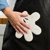 A hand rests on a large, glove-shaped pocket reminiscent of Mickey Mouse, set against black fabric—bringing a touch of Disney magic to the Apron with Glove Pockets.