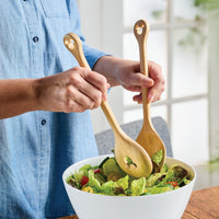 Using a 2-piece Bamboo Salad Server Set from the Disney Home Monochrome collection, a person mixes a fresh salad in a white bowl, adding a touch of whimsy with subtle Mickey Mouse designs.
