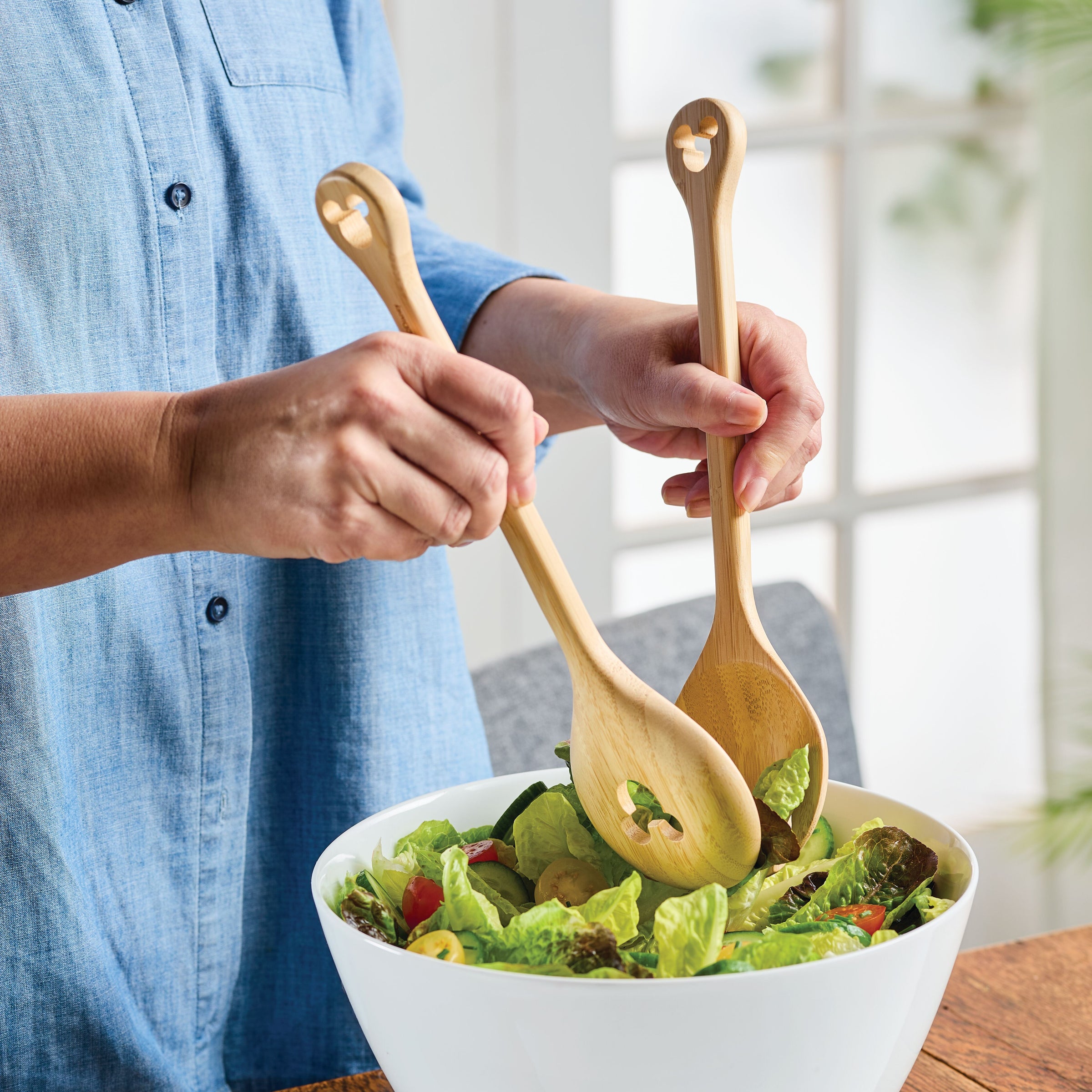 Using a 2-piece Bamboo Salad Server Set from the Disney Home Monochrome collection, a person mixes a fresh salad in a white bowl, adding a touch of whimsy with subtle Mickey Mouse designs.