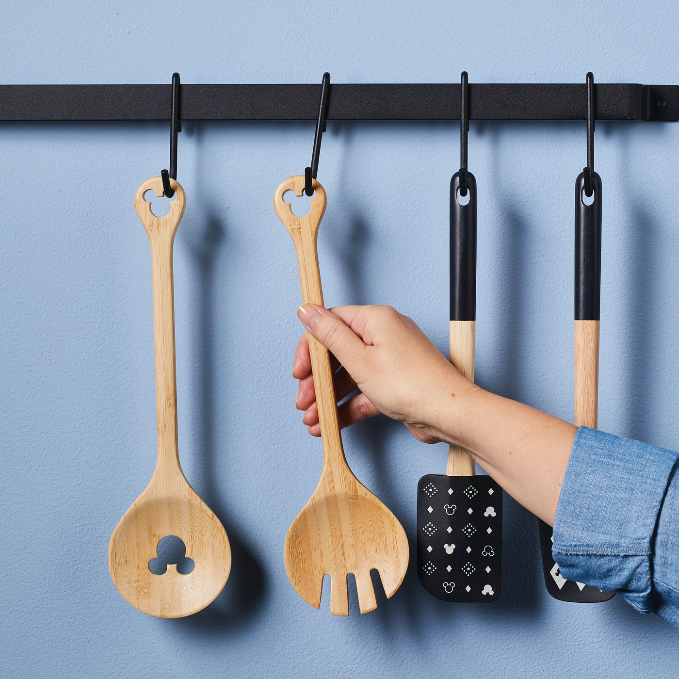 A hand reaches for a bamboo salad server from the hanging kitchen utensil rack, part of the charming Disney Home Monochrome collection.