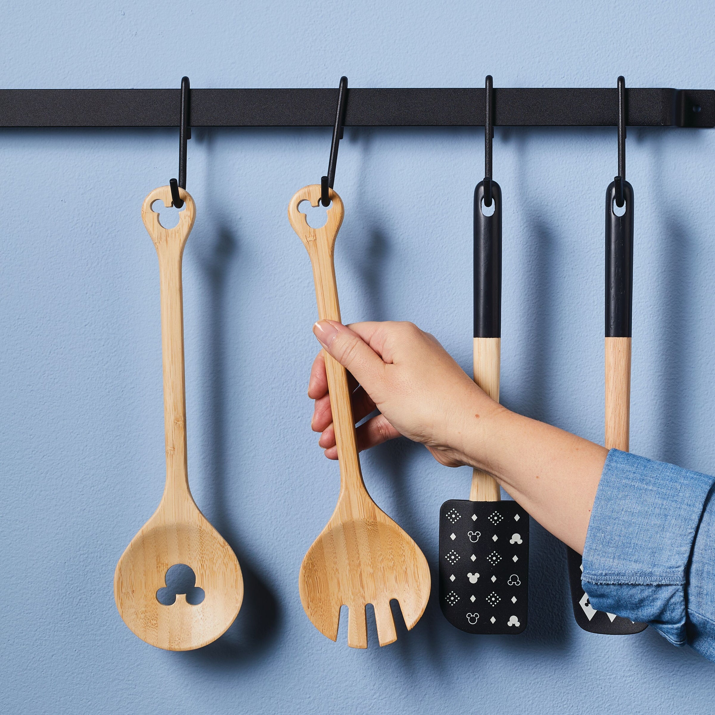 A hand reaches for a bamboo salad server from the hanging kitchen utensil rack, part of the charming Disney Home Monochrome collection.