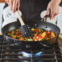A person is using the Monochrome Kitchen Tool Set to skillfully stir mixed vegetables in a frying pan on a gas stove.