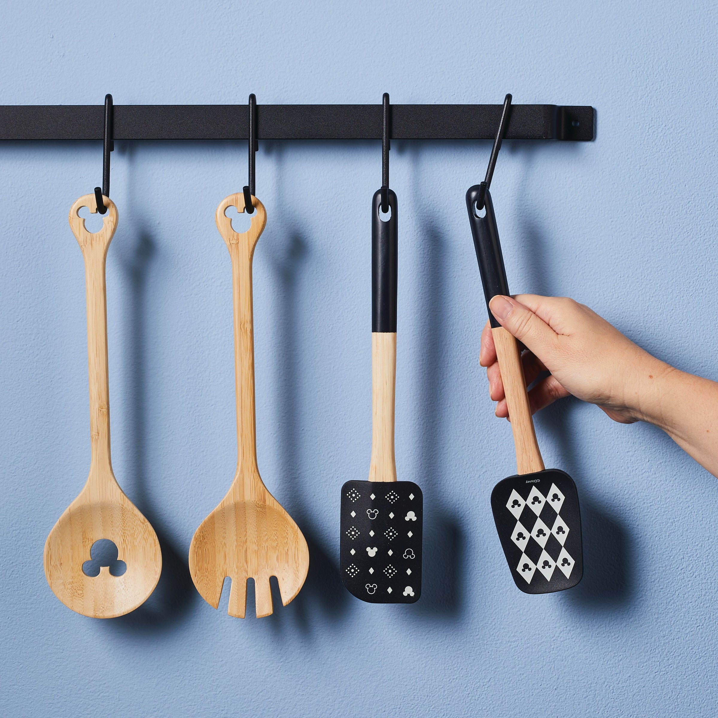 A hand holds a bamboo silicone spatula with a card suit design, as three utensils from the Monochrome Kitchen Tool Set hang on a rack, highlighting the charm of Disney kitchen tools.