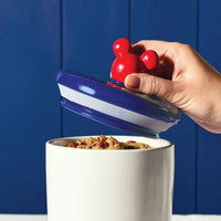 A hand lifts the lid with a red handle from the intricate Bon Voyage 2-Piece Ceramic Jar Set, showcasing its charming design.