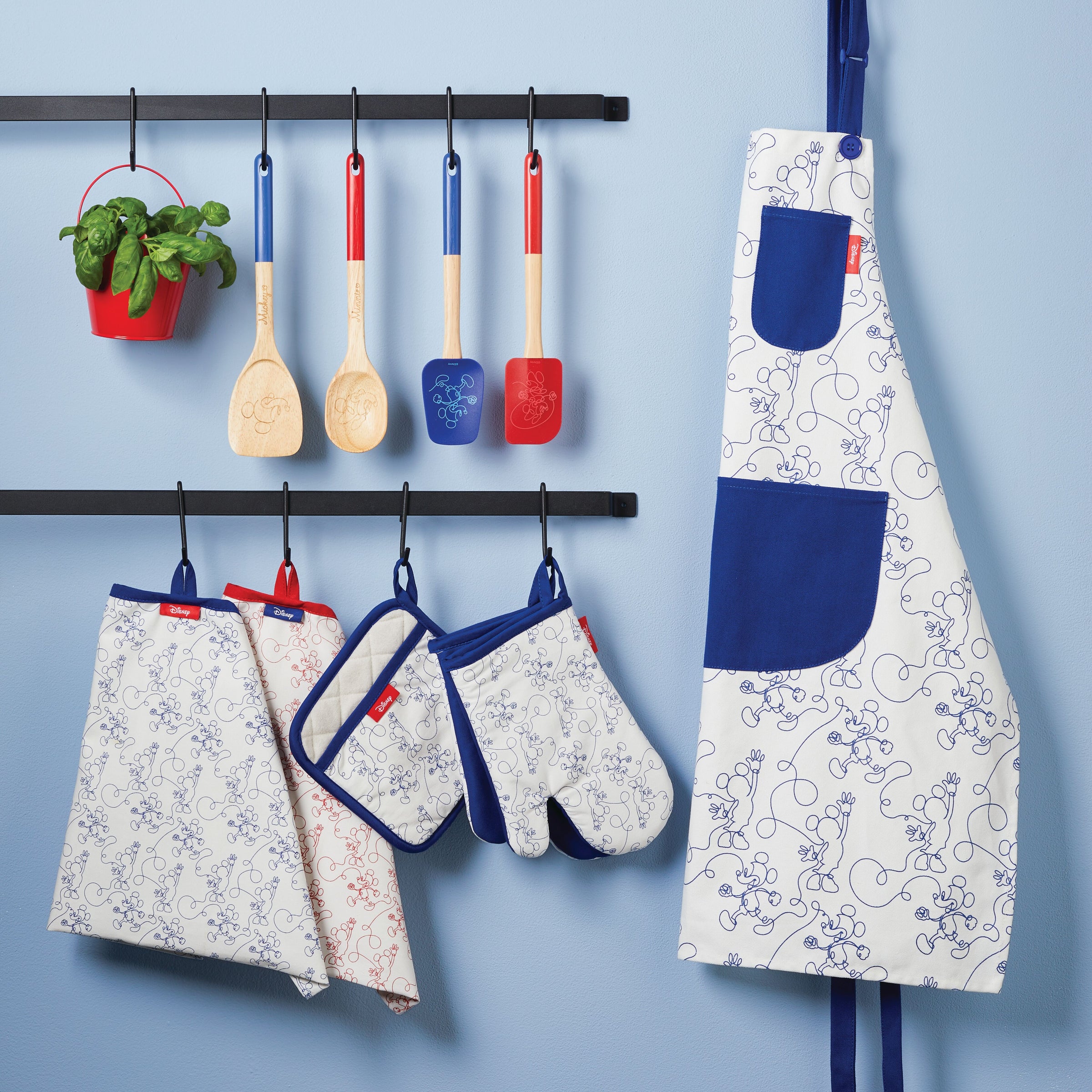 A kitchen apron set with utensils, featuring the Bon Voyage 2-Piece Spatula and Spoonula Set, hangs on a wall against a blue background.