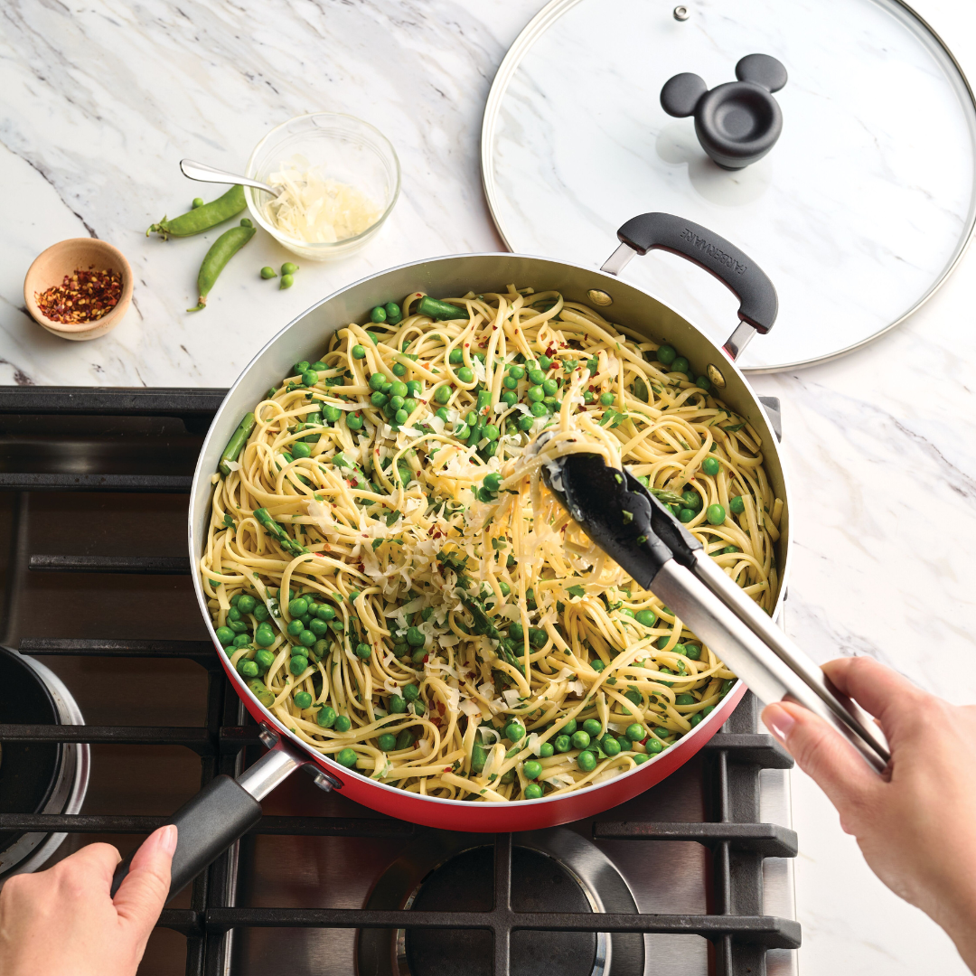 Person cooking spaghetti with peas in a red pan on a stovetop.