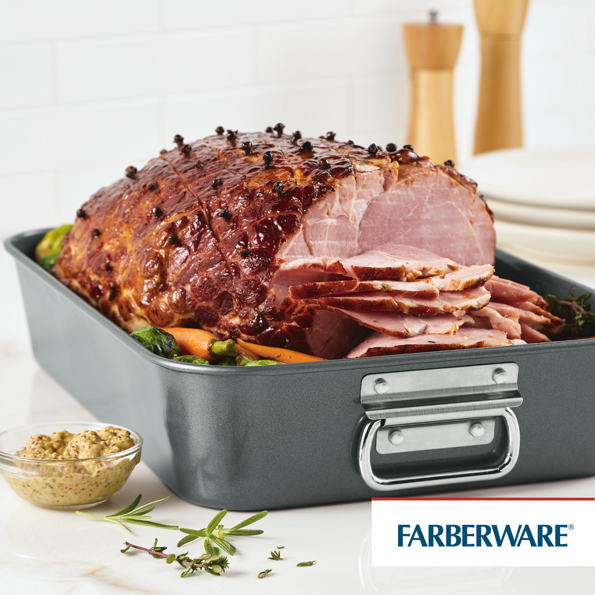 Sliced glazed ham in a roasting pan with vegetables and mustard on the side.