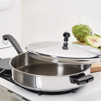 Stainless steel skillet with lid on a stovetop; artichokes in the background.