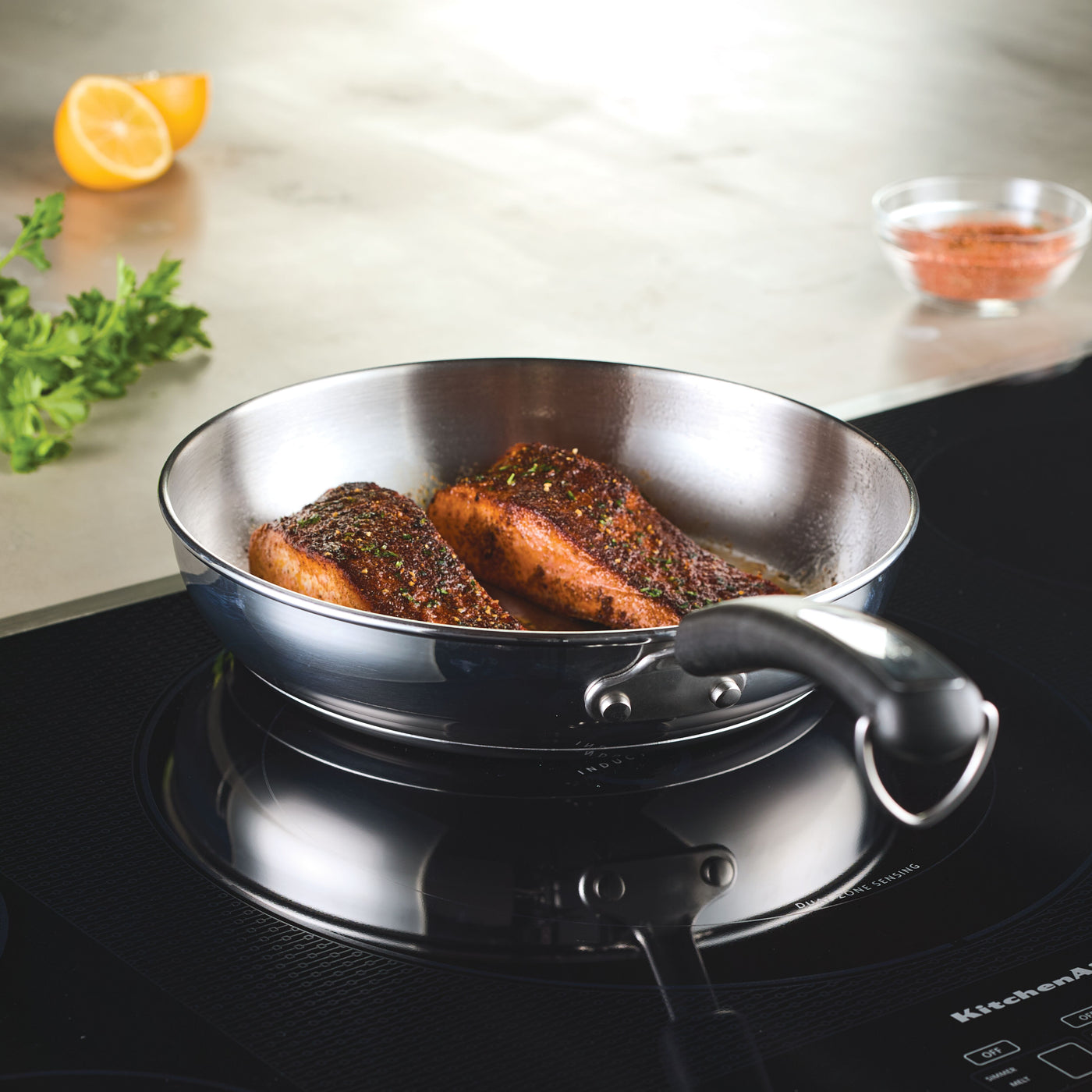 Salmon fillets sizzle in a skillet on the stovetop, accompanied by a vibrant display from the 10-Piece Stainless Steel Cookware Set, with fresh herbs and lemon enhancing the tantalizing aroma.
