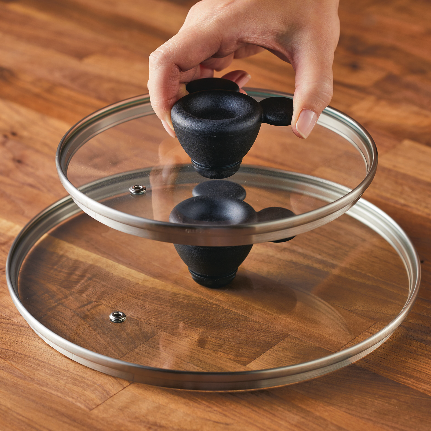 A hand holds a glass pot lid, its surface reflecting the warm tones of a wooden table, seamlessly fitting into the 7-Piece Monochrome Ceramic Nonstick Cookware Set.