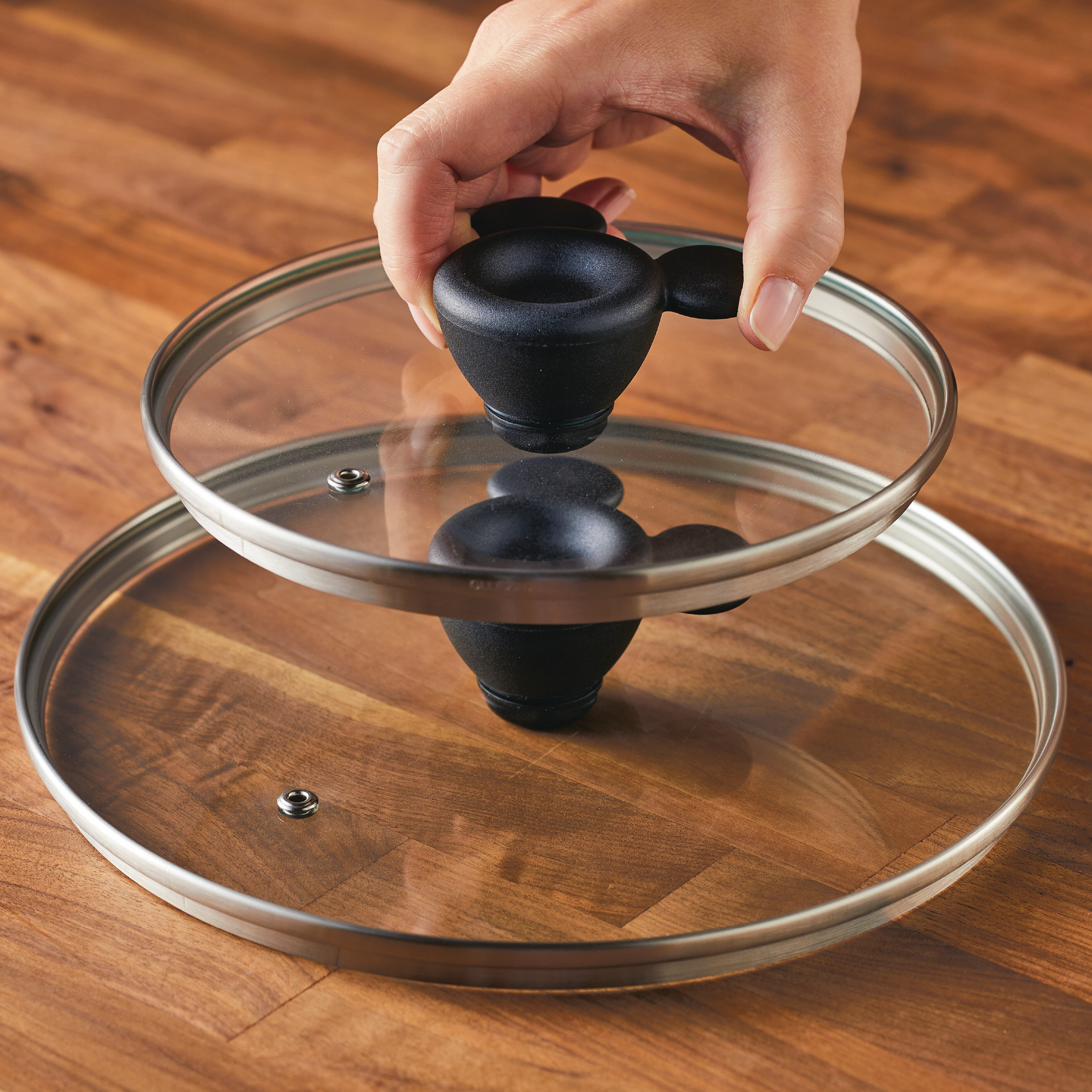 A hand holds a glass pot lid, its surface reflecting the warm tones of a wooden table, seamlessly fitting into the 7-Piece Monochrome Ceramic Nonstick Cookware Set.