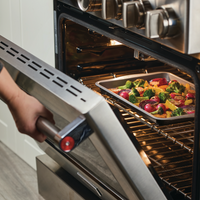A person is slipping a tray of mixed vegetables into the open oven, using their favorite 10" x 15" Nonstick Cookie Pan.