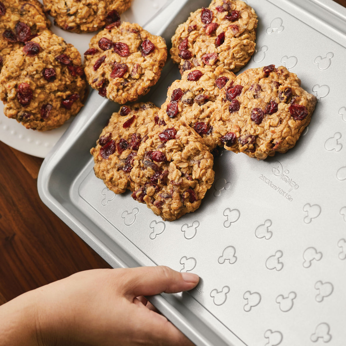 Hand placing oatmeal cookies with cranberries on a Mickey-design baking sheet.