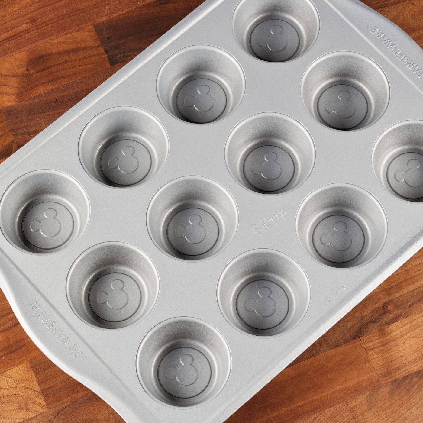 A 12-cup nonstick muffin pan, designed with whimsical Mickey Mouse-shaped molds, sits on a wooden surface—an ideal addition to any Disney-themed bakeware collection.