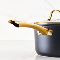 A gold-handled black pot with a glass lid from the Forged Induction Ceramic 12-Piece Cookware Set rests elegantly on a marble surface, offering versatility for induction cooking.