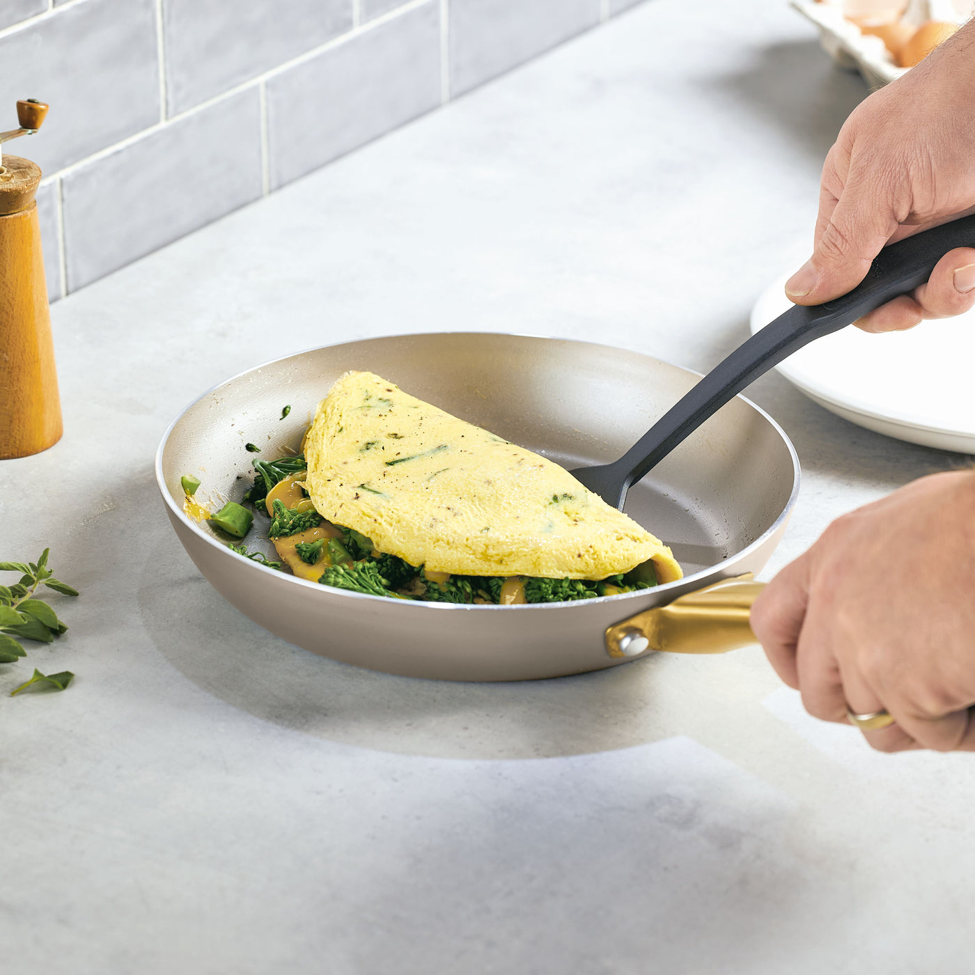 A person expertly folds an omelet with spinach in a frying pan from the Radiant Nonstick 12-Piece Cookware Set, appreciating its even heat distribution. The gold accent handle adds a touch of elegance as they create a perfectly crafted breakfast.