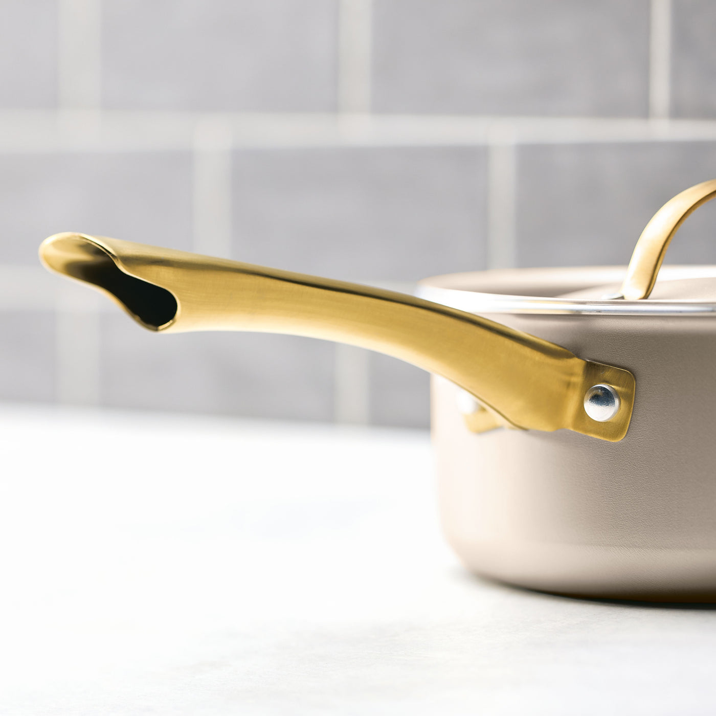 Close-up of the Radiant Nonstick 2-Quart Saucepan With Lid in beige, featuring a gold handle, placed on a kitchen counter and designed for even heat distribution.