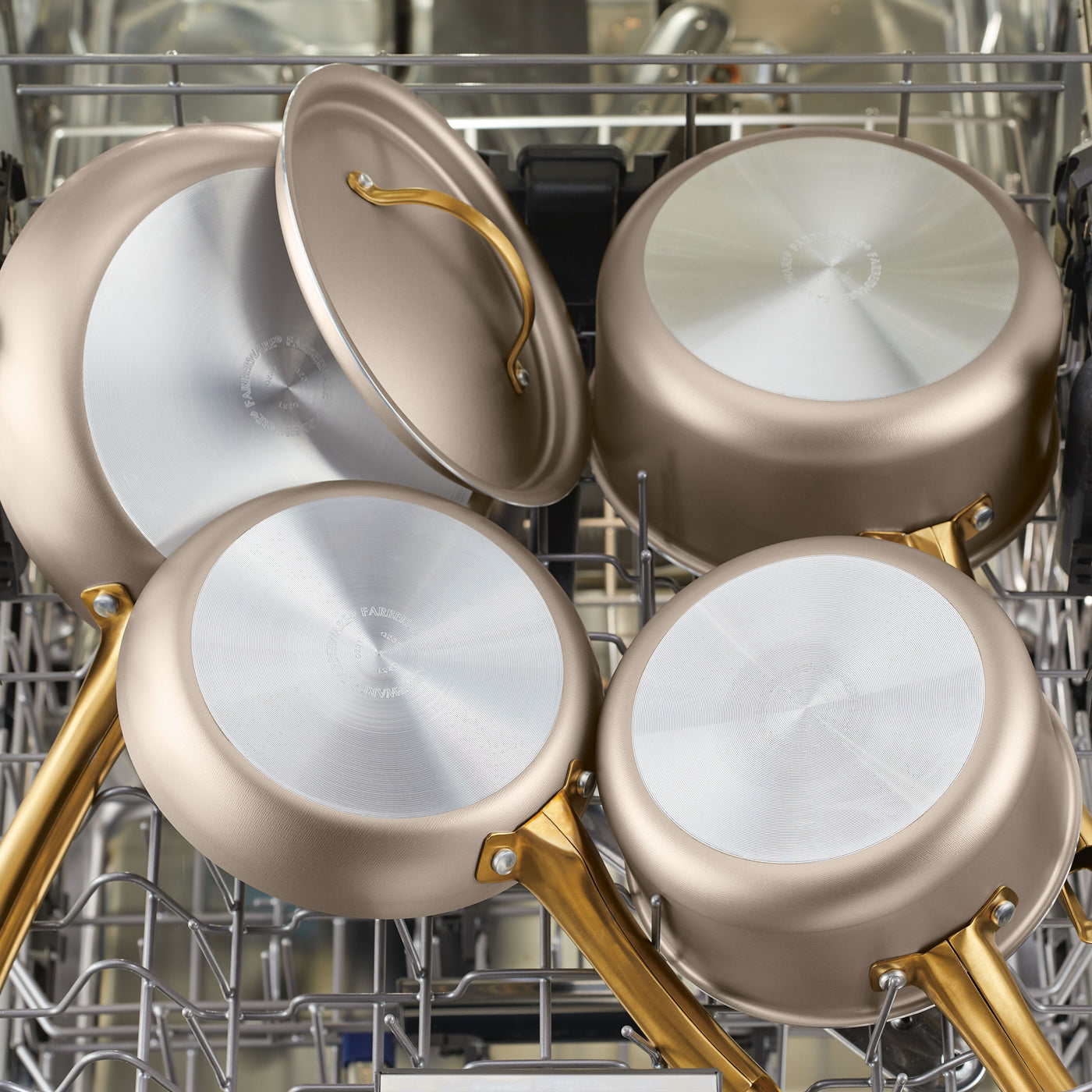 Four Radiant Nonstick Frying Pans, designed with silver bodies and gold handles for even heat distribution and featuring a champagne-colored surface, are placed in a dishwasher.