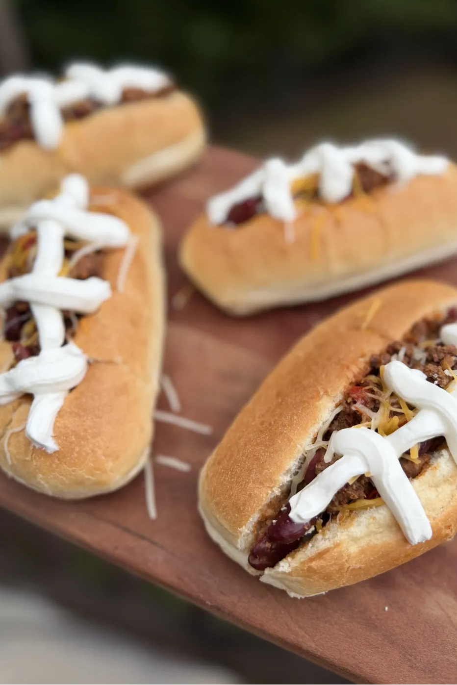 Four mini football-style hot dogs on a wooden serving board, topped with melted cheese and sour cream patterned like laces, with a blurred outdoor background.