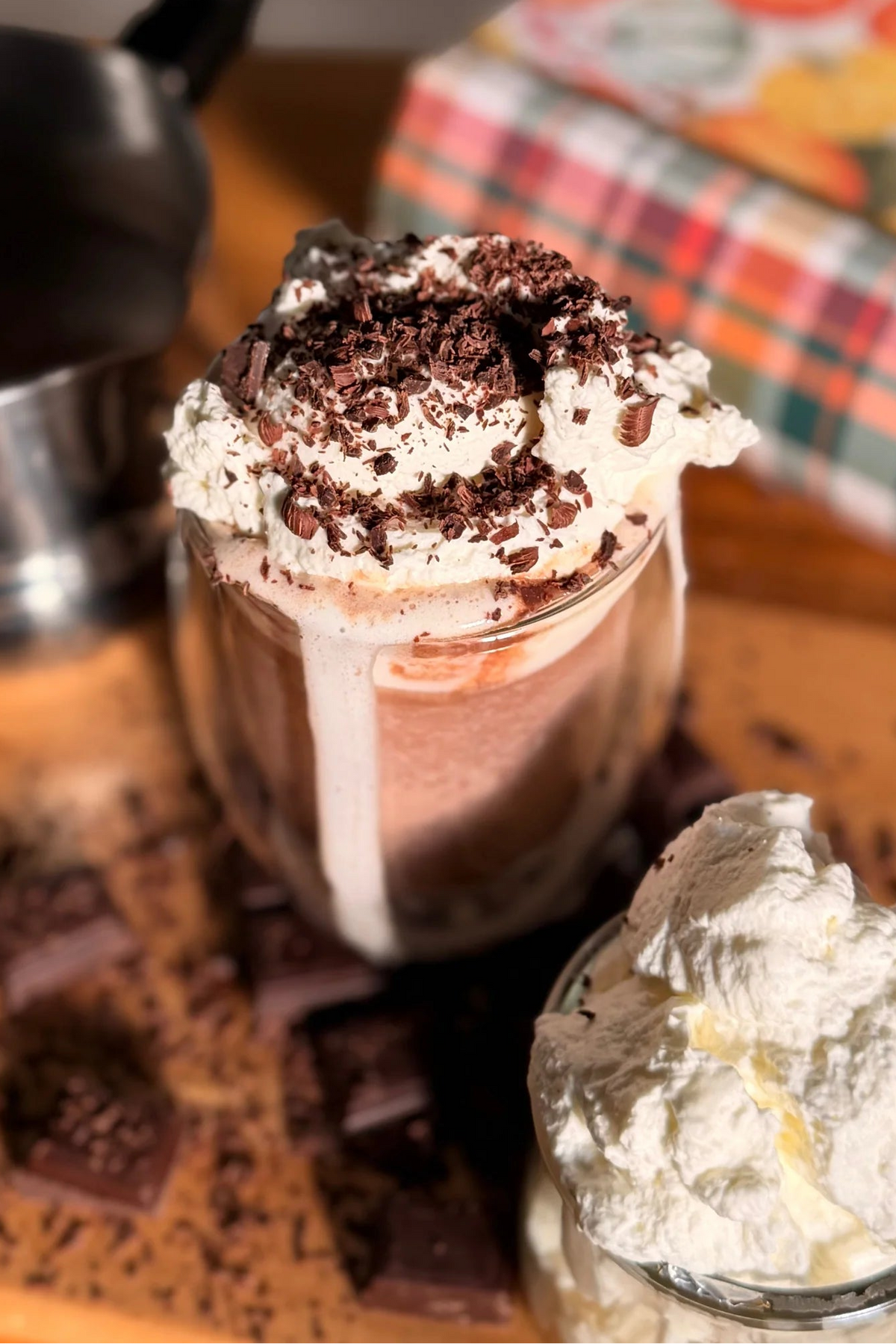A hot chocolate or cocoa drink in a clear glass jar topped with whipped cream and chocolate crumbs, shown on a wooden surface with chocolate pieces, a small bowl of whipped cream, and a blurred plaid cloth in the background.