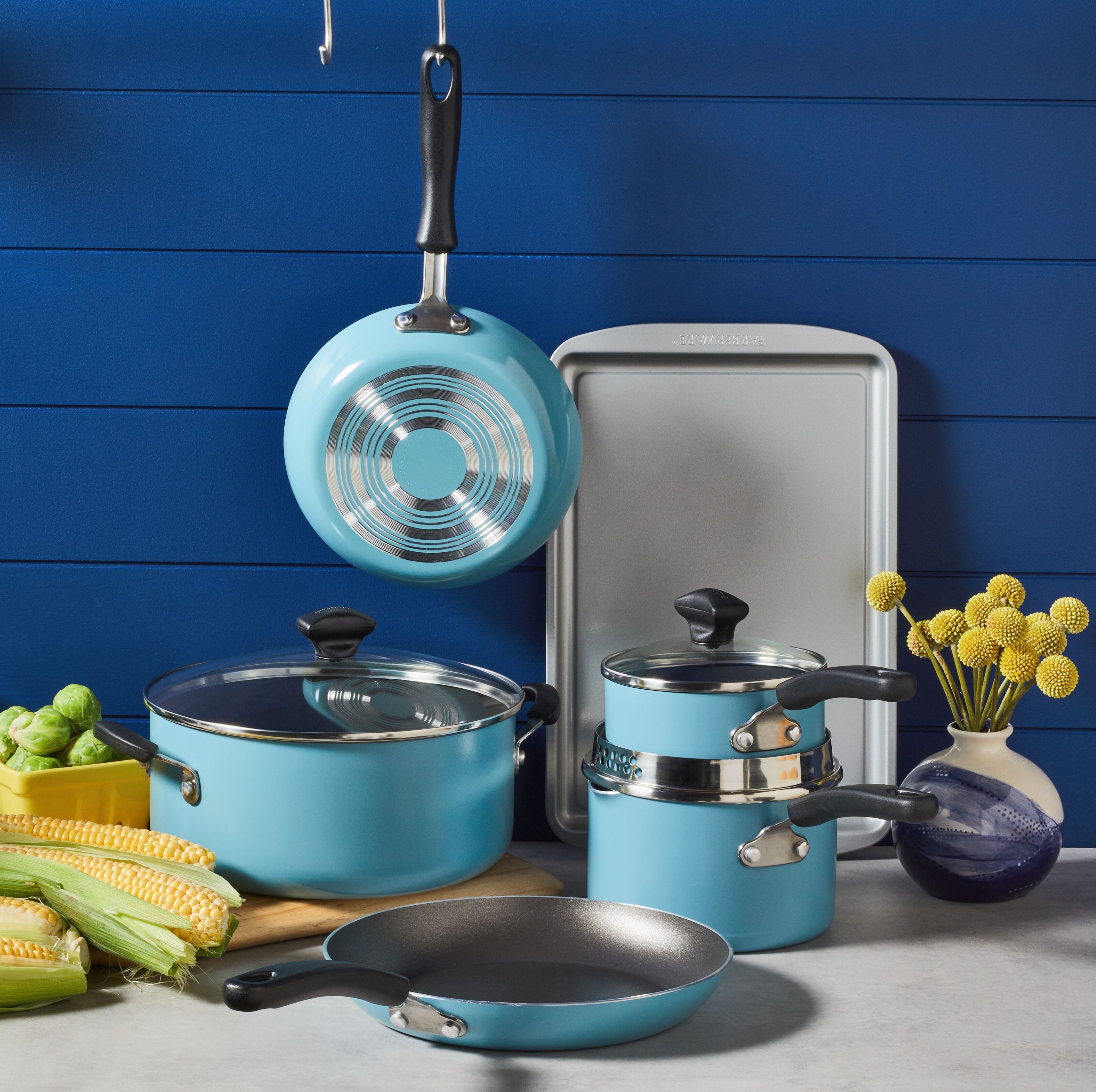 A 15-Piece Nonstick Cookware Set in teal, made from heavy gauge aluminum, is complemented by corn, Brussels sprouts, and decor on a counter.