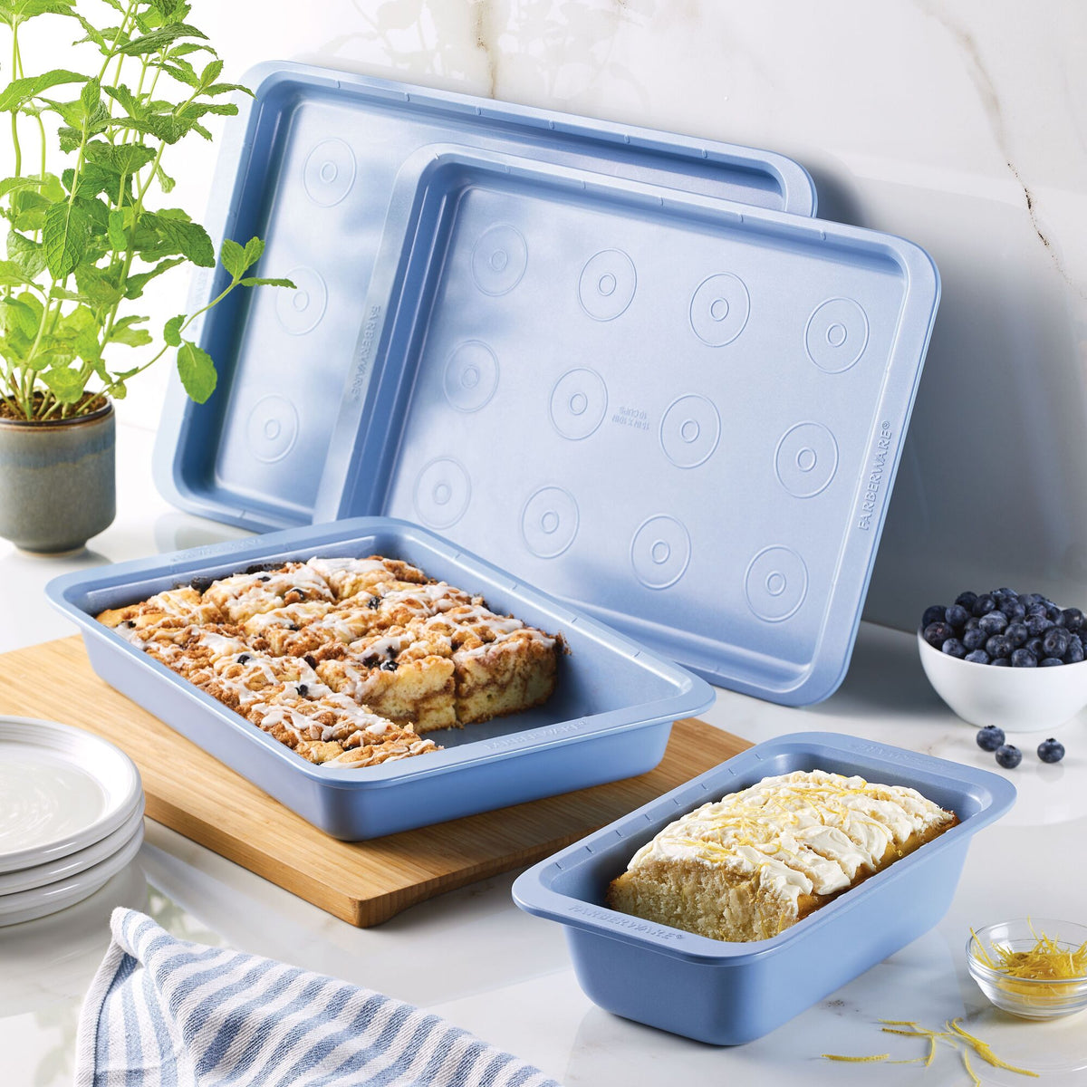 Blue baking pans with desserts beside a plant and plates on a marble counter.