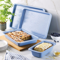 Blue baking pans with desserts beside a plant and plates on a marble counter.