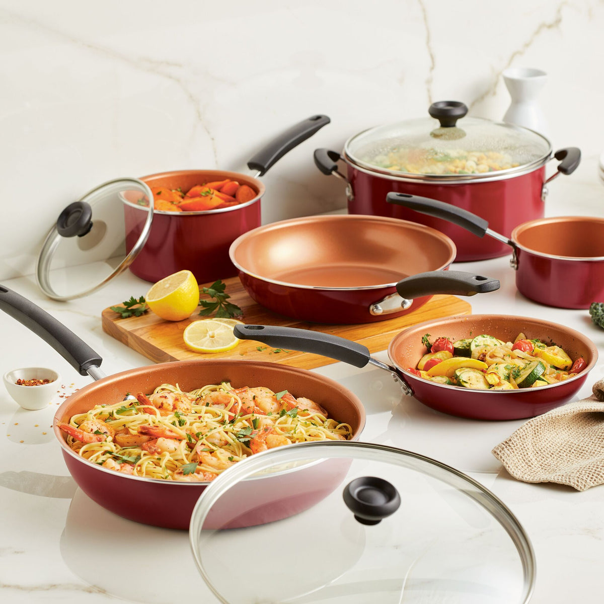 Red cookware set with food, lemon, and glass lids on a marble countertop.