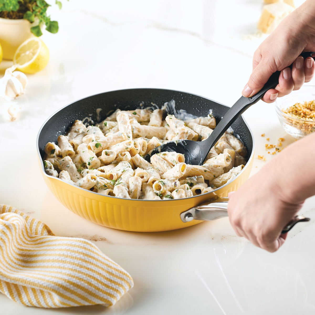 Hands stirring creamy pasta in a yellow pan with a black spoon.