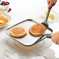 Two pancakes being flipped on a square pan with a spatula.