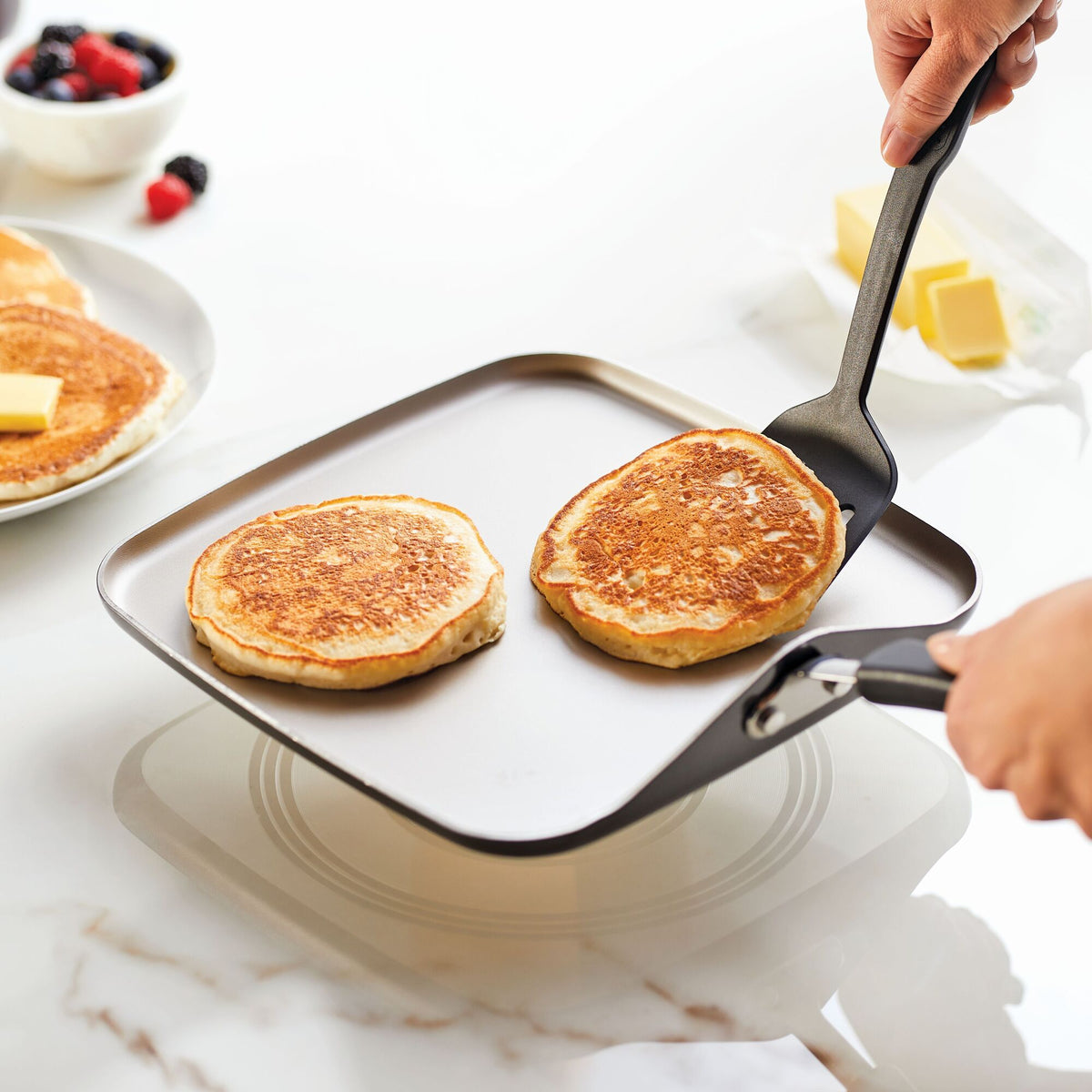 Two pancakes being flipped on a square pan with a spatula.