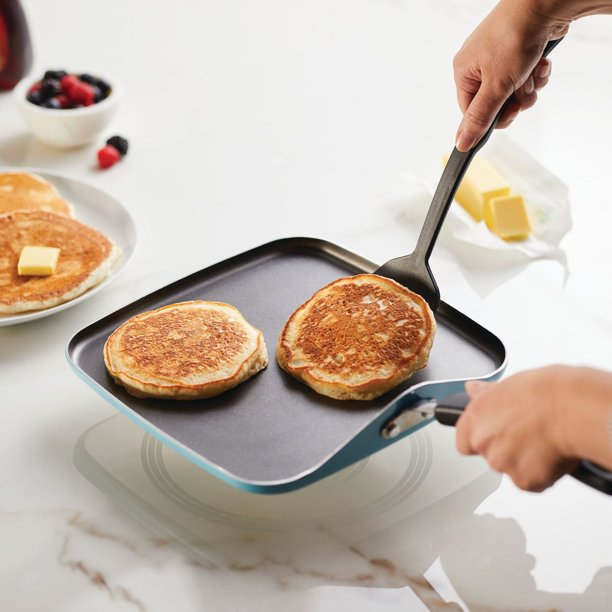 Person cooking pancakes on a griddle with butter and fruit nearby.