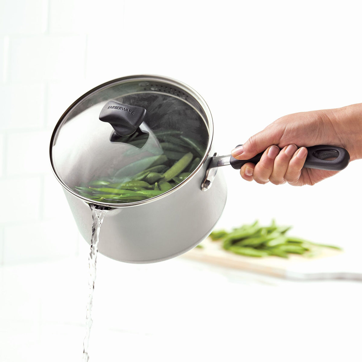 A person drains water from a pot with a lid, holding green beans inside.