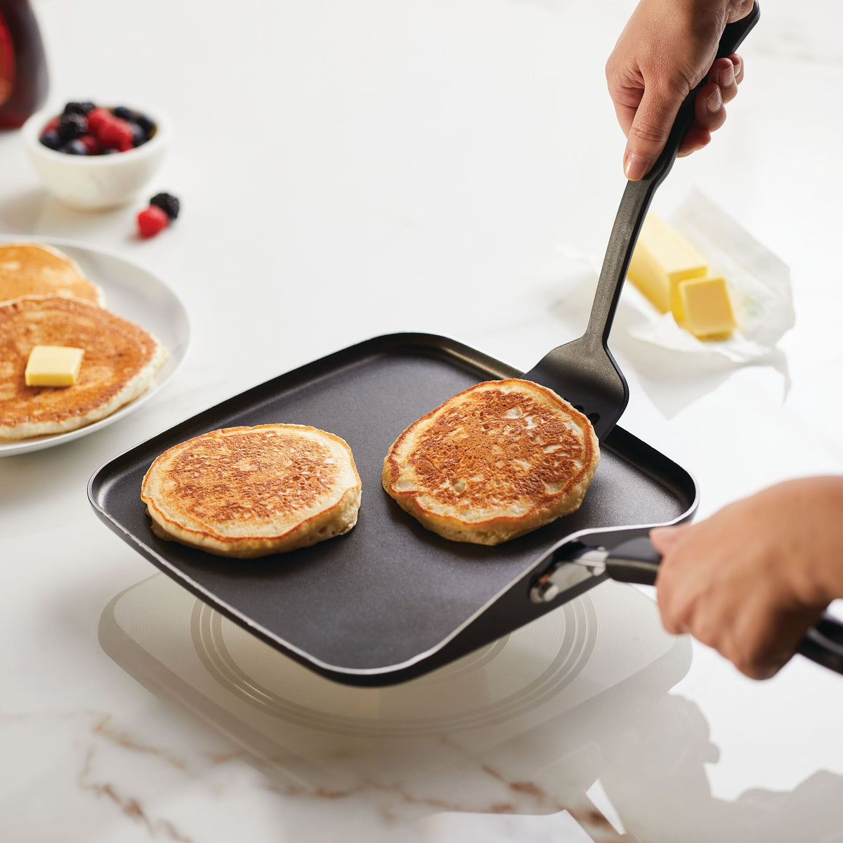 Two pancakes being flipped in a square pan on a stovetop.