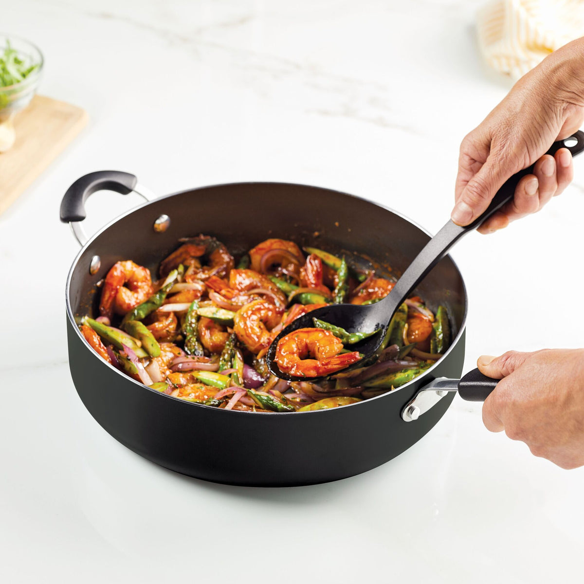 Hands stirring shrimp and vegetables in a black skillet on a stove.