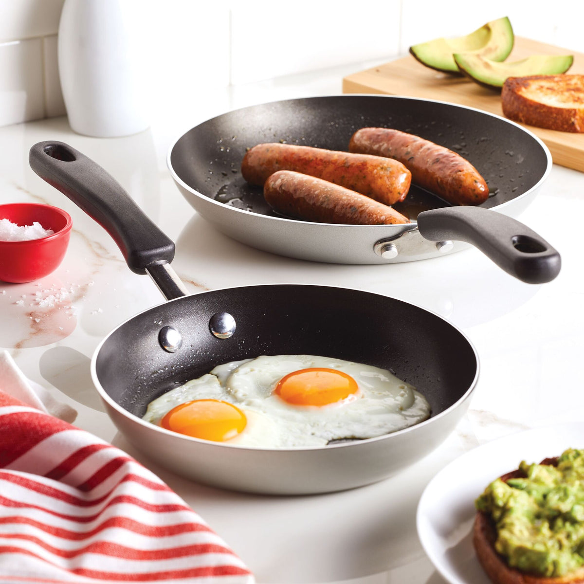 Two frying pans on a stove, one with eggs and the other with sausages.