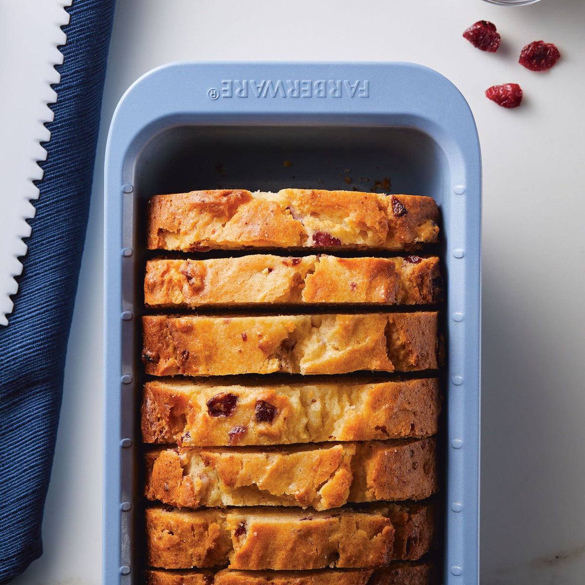 Sliced cranberry loaf cake in a blue loaf pan on a white surface.