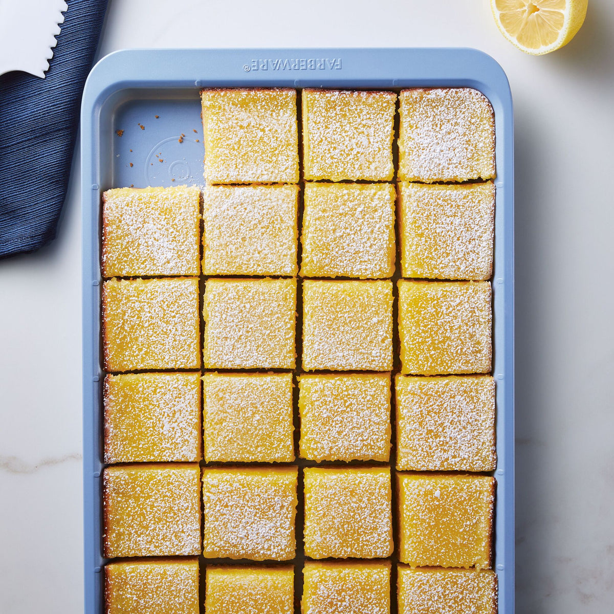 A tray of lemon bars dusted with powdered sugar, next to a lemon half.