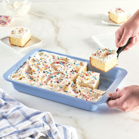 Hands serving a frosted cake with sprinkles from a blue baking dish.