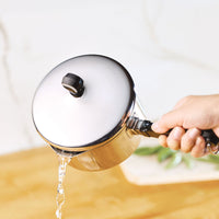 Hand pouring water from a lidded saucepan on a wooden surface.