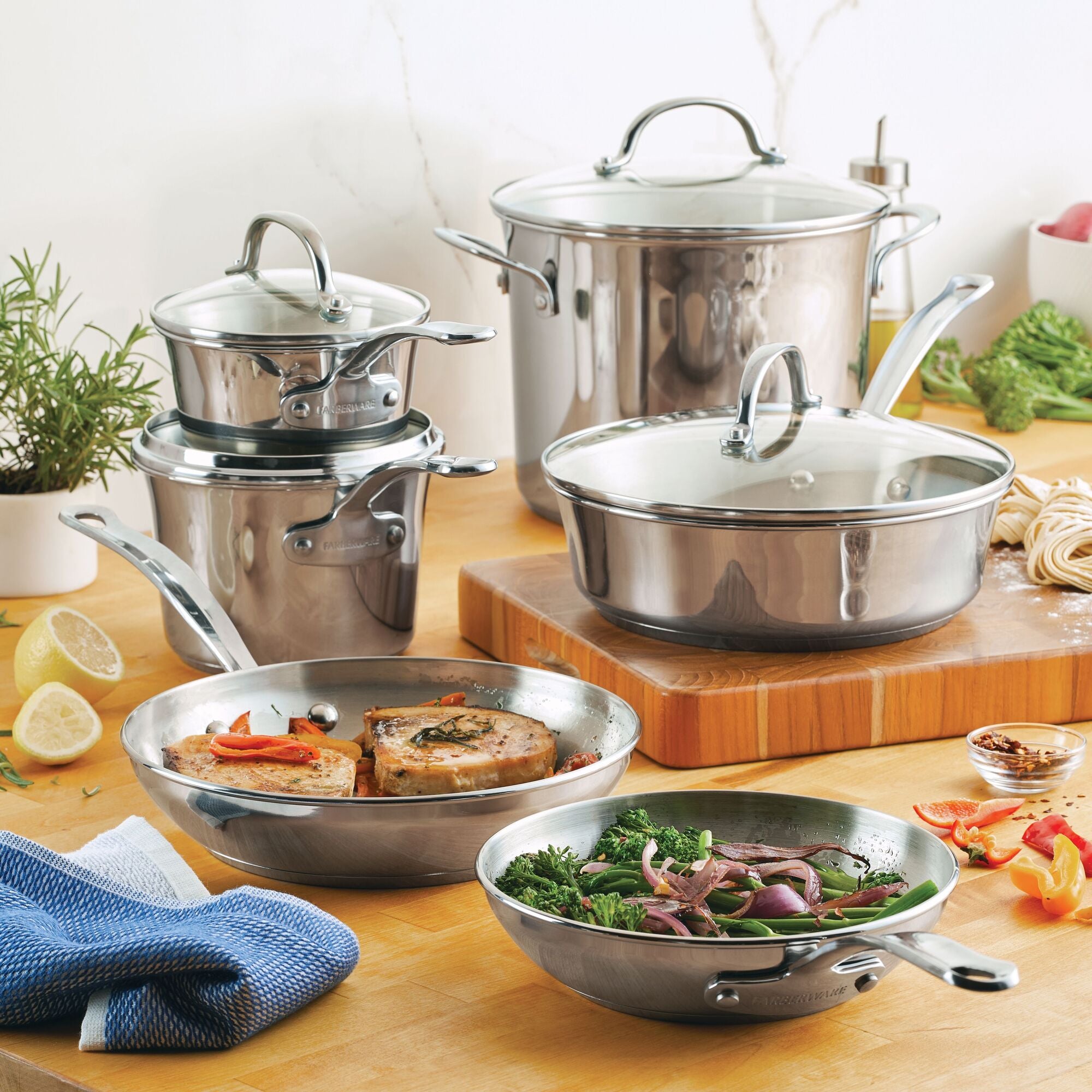 The Millennium Stainless Steel 10-Piece Cookware Set sits on a wooden kitchen counter, surrounded by fresh vegetables. Known for even heat distribution and being dishwasher safe, these pieces are both practical and stylish for any kitchen.
